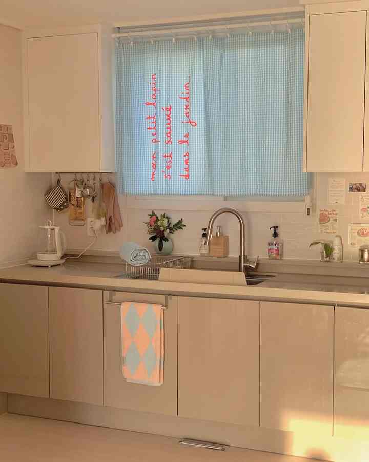 Pastel blue curtain and modern beige kitchen cabinets with neatly arranged kitchen utensils create a cozy, clean kitchen space