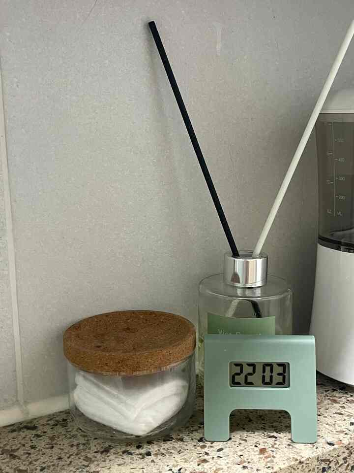 Bathroom shelf with soft natural tones featuring a small olive-colored digital clock, cork lid glass container, and diffuser creating a cozy atmosphere
