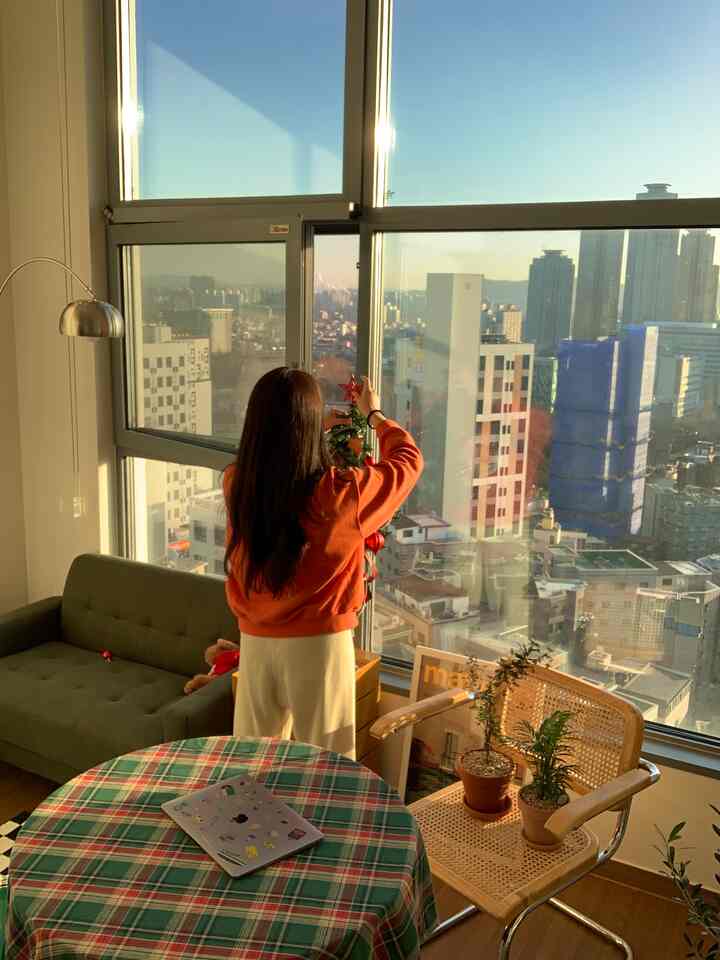 A cozy studio apartment with natural warm tones, featuring a Christmas tree by the window and a plaid tablecloth on a round table holding a laptop in a home office setup