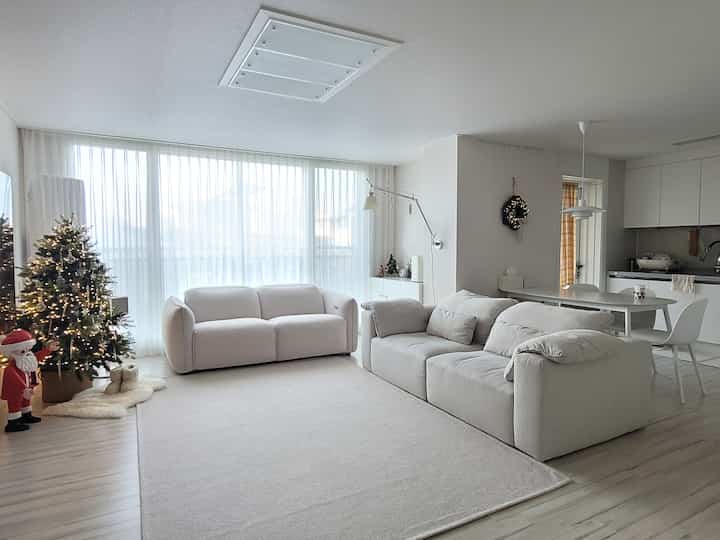 White and beige toned living and dining room with two sofas, Christmas tree, Santa figure, and wreath lighting creating a cozy winter atmosphere