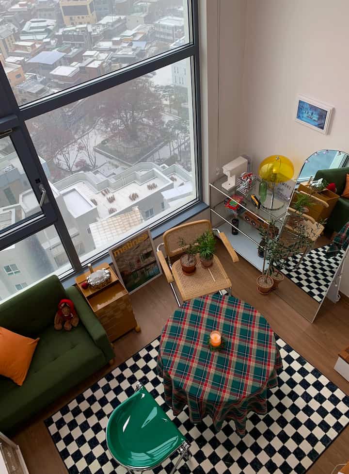 Green and beige toned studio living room featuring a checkered rug, round table, vintage-style furniture and warm lighting creating a cozy atmosphere