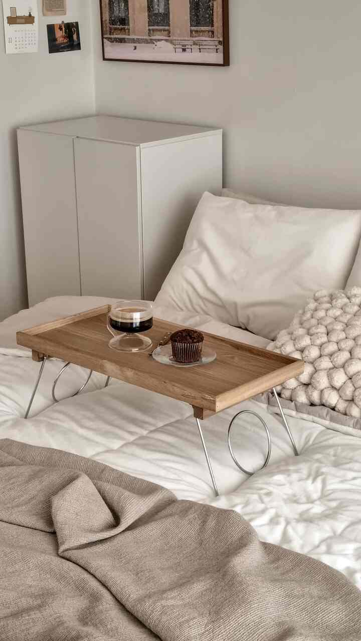 A cozy bedroom in white and brown tones featuring a wooden tray with Americano and chocolate muffin on the bed
