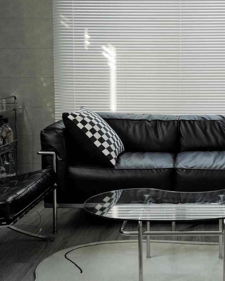 Modern living room featuring black leather sofa with checkered cushion, white blinds, and glass coffee table