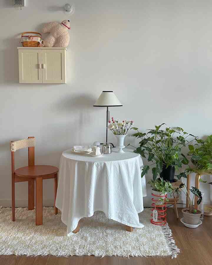 Natural-toned small entrance area featuring a wood stool, round table with white tablecloth, plants, and lamp in a cozy atmosphere