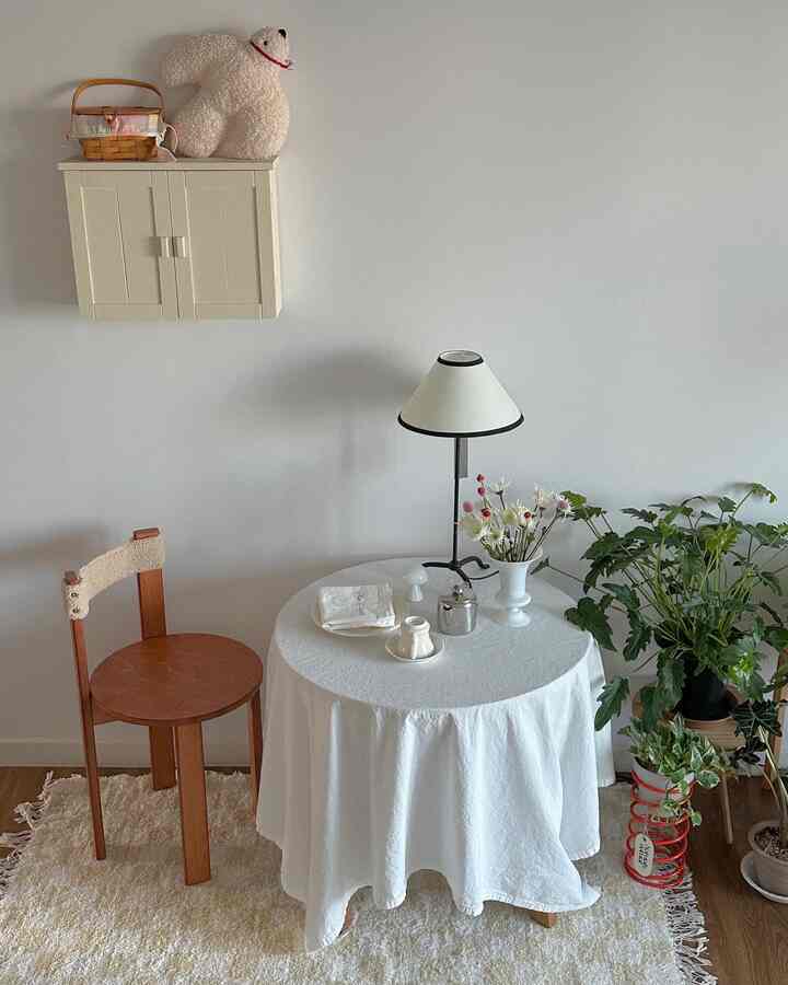 ホワイトとナチュラルトーンの小さなリビング、木製のダイニングチェアと白いテーブルクロスがかかった丸テーブル、多彩な植物とフロアランプが調和した居心地の良い空間