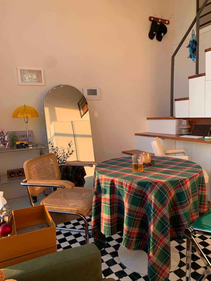Warm natural-toned studio apartment featuring a table with Christmas plaid tablecloth, rattan and white chairs for a cozy home party setup