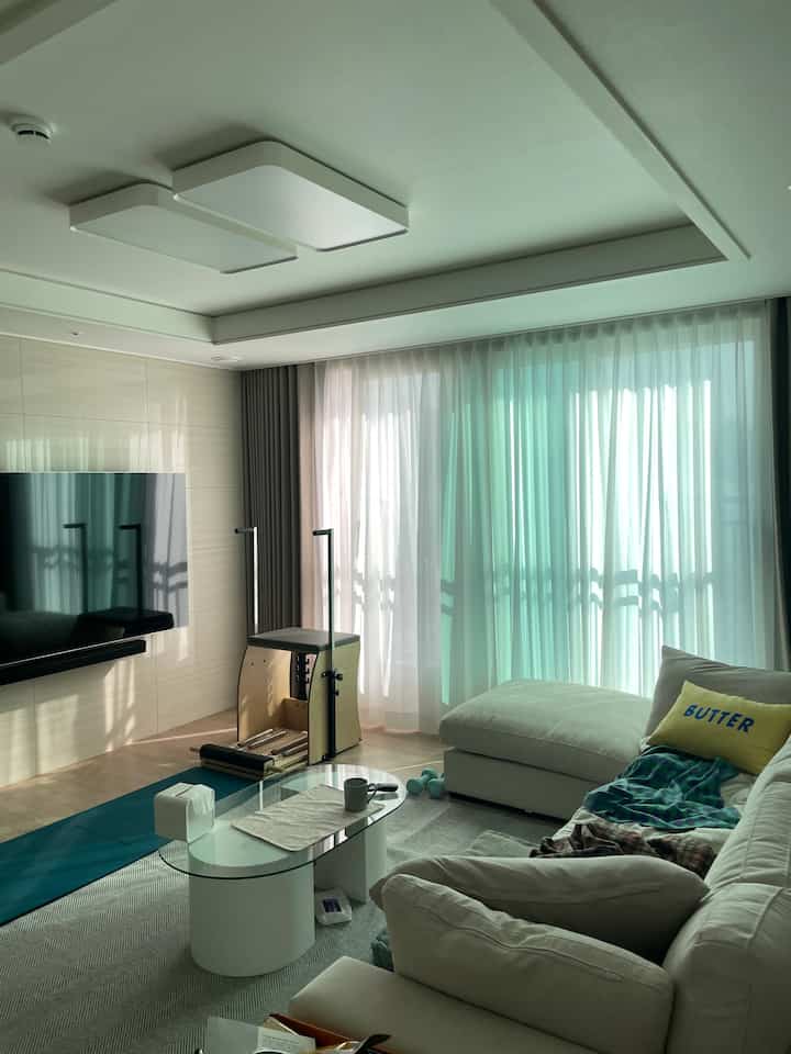 Natural-toned living room with large fabric sofa, round glass coffee table, Pilates equipment, and sheer plus blackout curtains