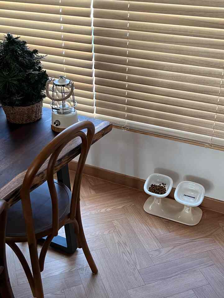 Wood tone corner of residential space featuring wooden blinds, dining table and chair, with adjustable-height cat feeding bowls on the floor