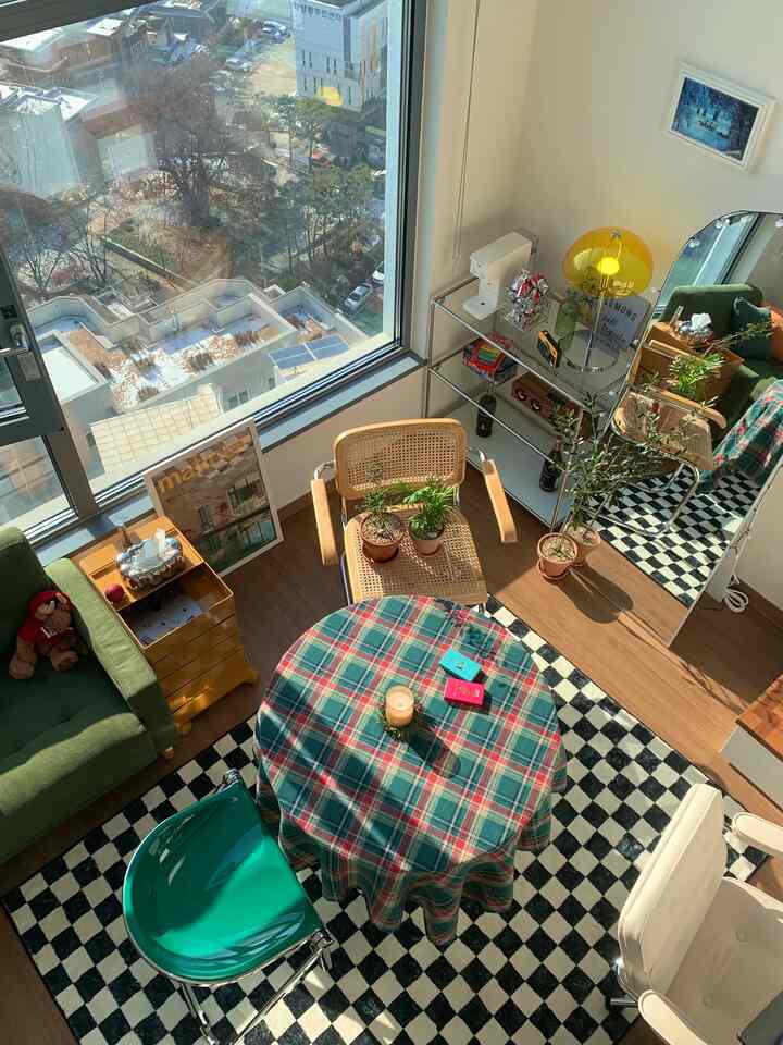 白い壁と木目調の床のワンルームリビング、ヴィンテージ家具と緑のソファ、チェック柄のテーブルクロスが配置された雰囲気のあるインテリア