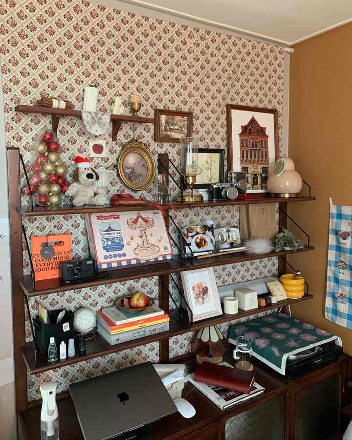 A cozy small room with vintage patterned wallpaper and wooden wall shelves, decorated with Christmas ornaments and various objets