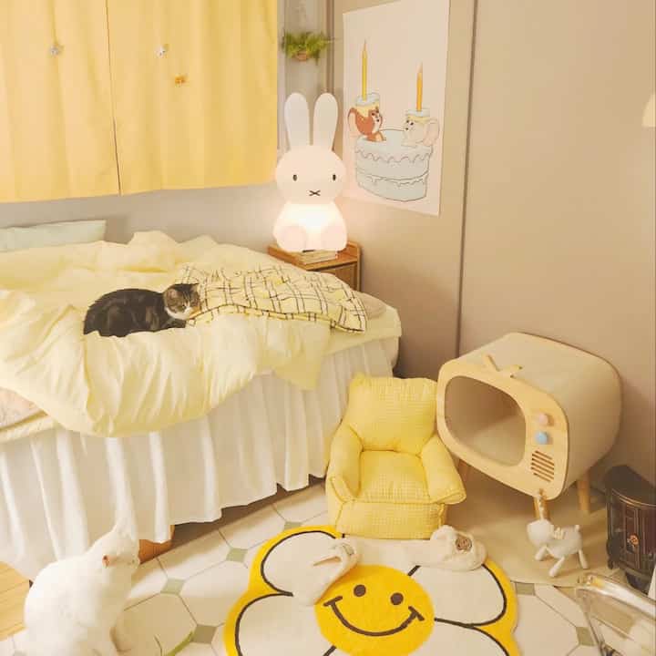 Warm yellow-toned bedroom with cats resting on the duvet and floor, featuring a bunny-shaped lamp and cozy decorative elements