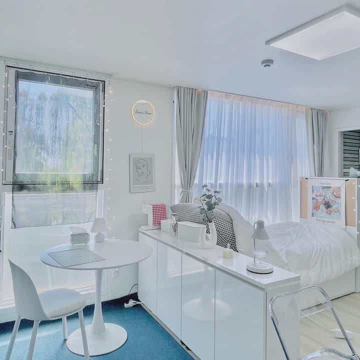 Bright white-toned studio apartment featuring a white dining table and chair, bed, and long storage cabinet in a clean and airy space