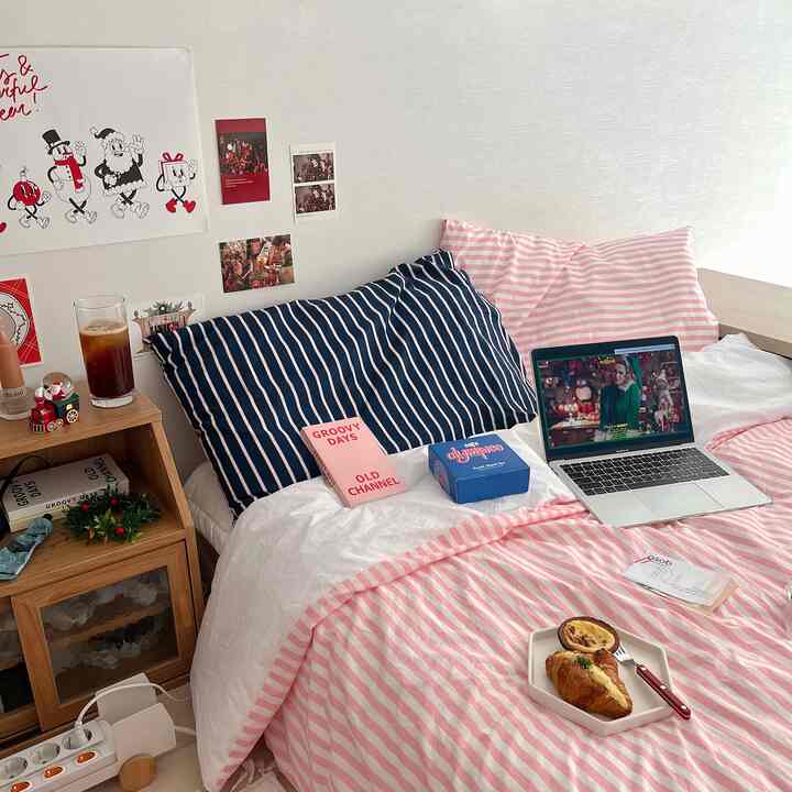 白い壁とピンクのストライプ柄布団カバーが調和した居心地の良い寝室、ノートパソコンとコーヒーが置かれたおうちカフェの雰囲気のあるインテリア