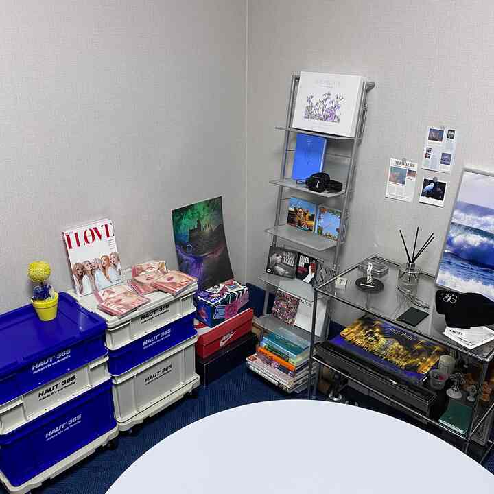 青いカーペットと白い収納ケースが配置された一人暮らし向けの狭い部屋、多目的な本棚とガラスデスクがある印象的な空間