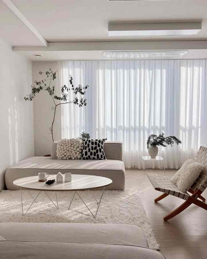 A bright living room with natural colors and wood tones, featuring a large sofa, coffee table, plants, and soft curtains creating a cozy atmosphere.