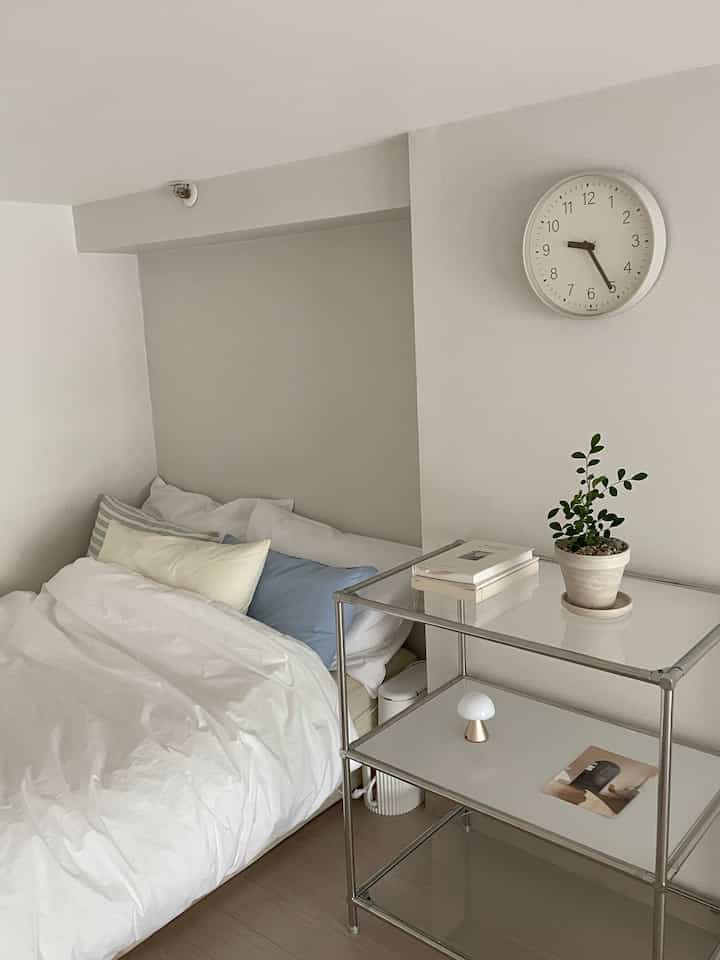 ホワイトとナチュラルの色調が調和したミニマルな寝室、清潔感のある寝具と棚、時計が配置された雰囲気のあるインテリア