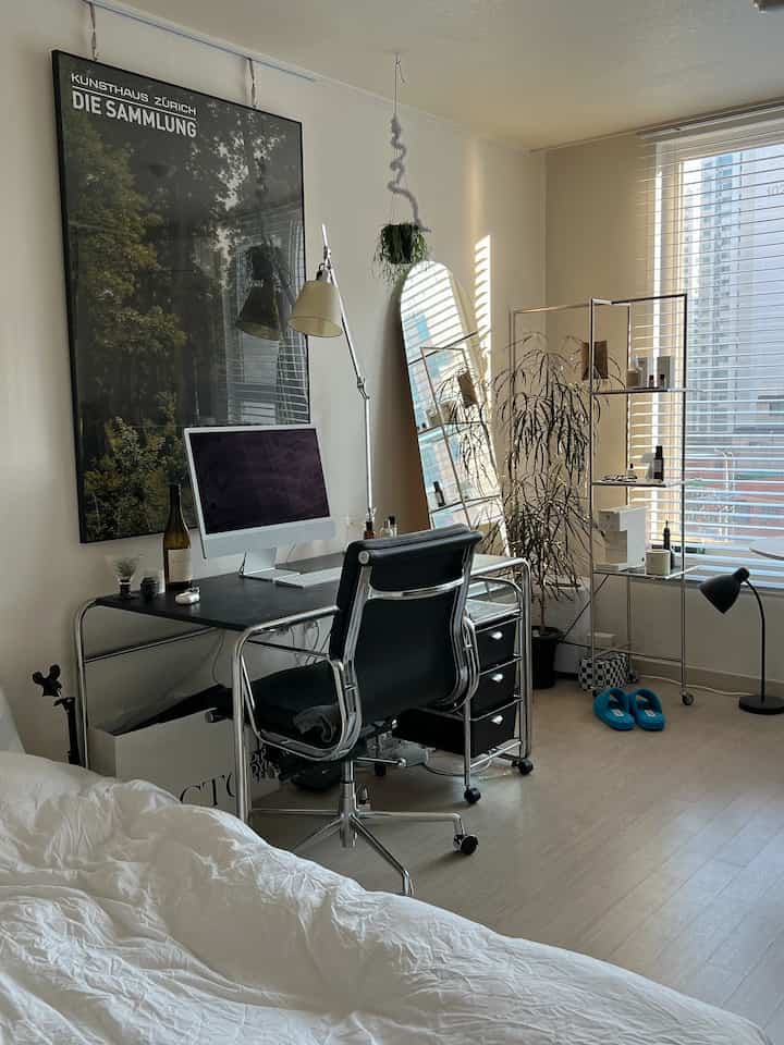 Bright white and black toned single household bedroom with a home office setup, featuring natural light from windows and indoor plants