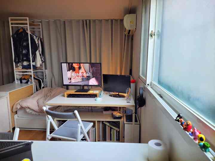 White and brown toned small home office and bedroom space featuring clean desk, bed, and gray curtains background