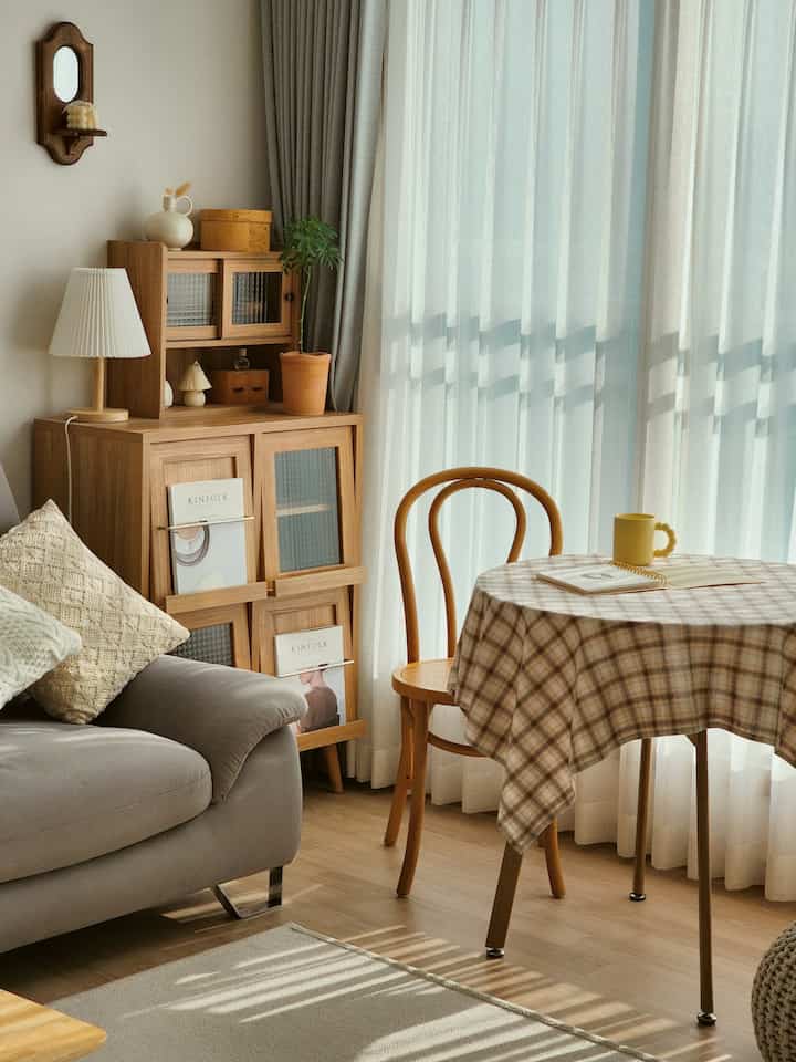 A cozy living room with natural beige and wood tones, featuring a wooden cabinet, checkered table, and soft-lit lace curtains