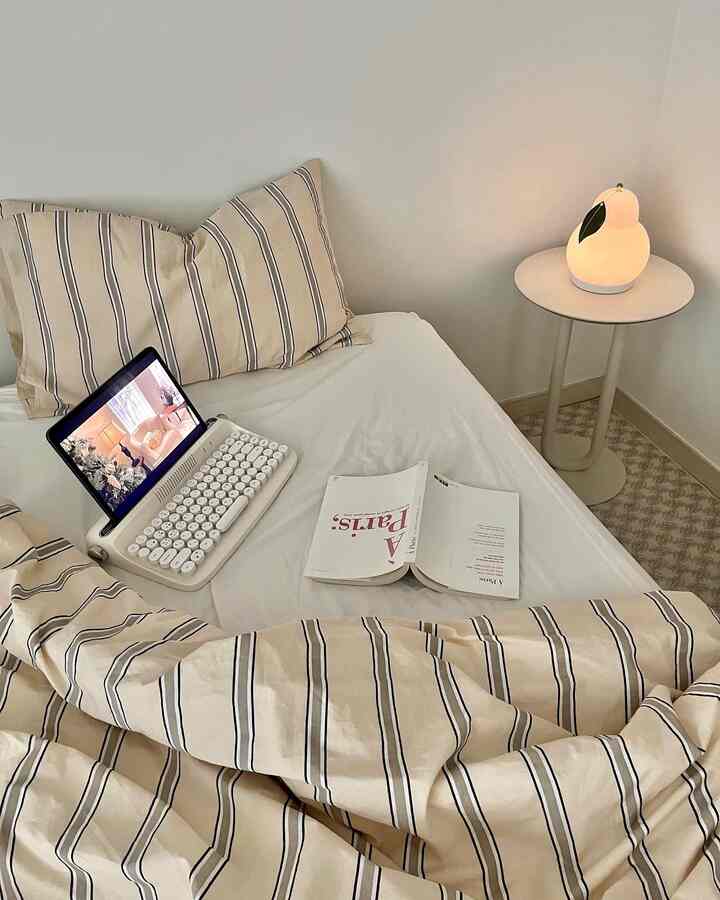 A beige and white toned bedroom featuring a striped bedding bed centered and a nightstand with a lamp on the right, creating a cozy atmosphere