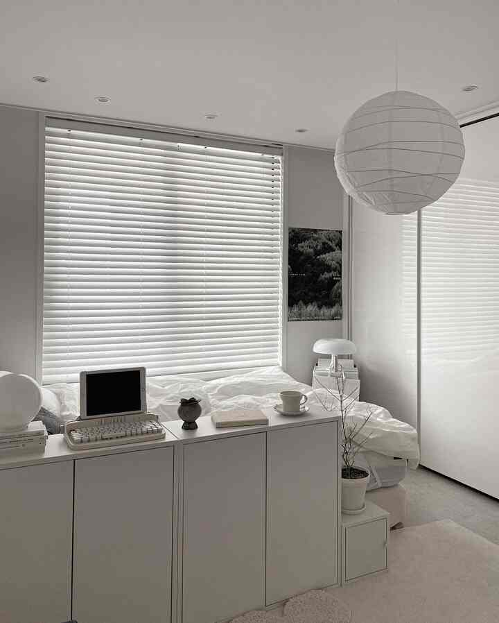 Minimal white-tone bedroom featuring blinds on window, neat furniture arrangement, and natural accents