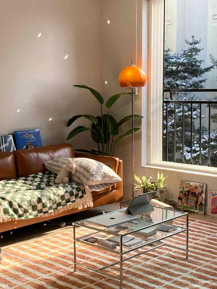 明るく暖かみのあるブラウンのリビング、レザーソファとガラスのコーヒーテーブル、オレンジのフロアランプが配置された自然光あふれる空間