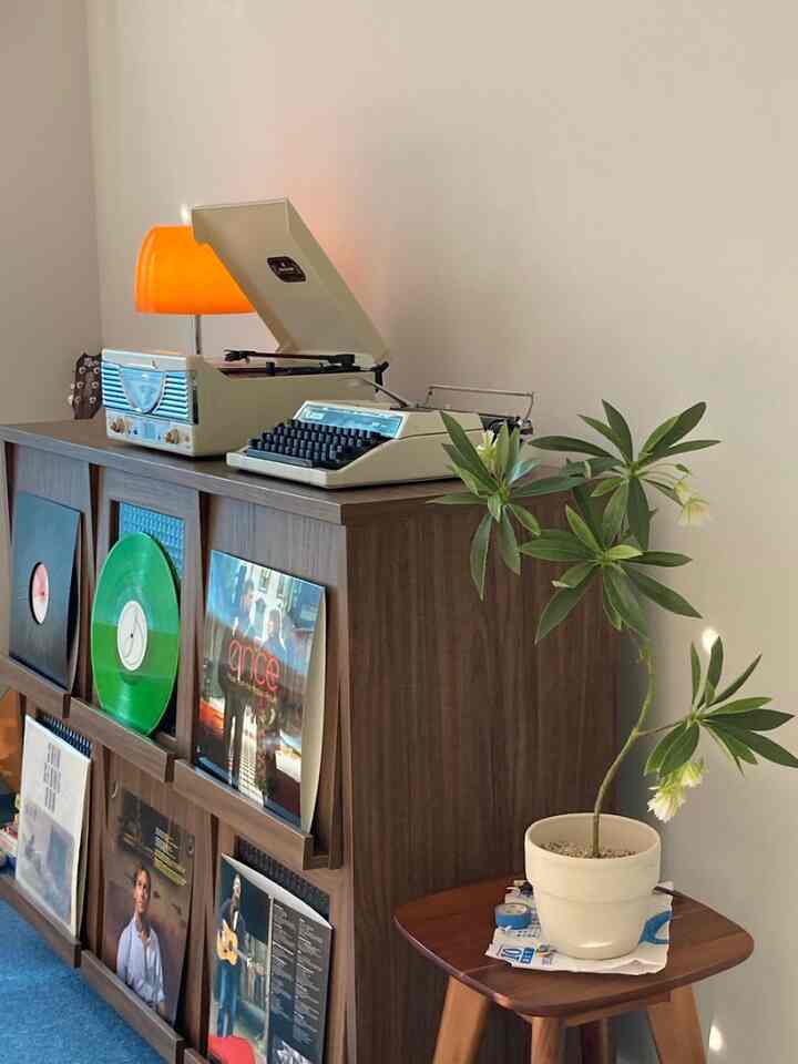 レトロな雰囲気のリビング空間、木目調キャビネットの上にヴィンテージのタイプライターとレコードプレーヤーが置かれ、スツールの上に観葉植物が配置された温かみのあるインテリア