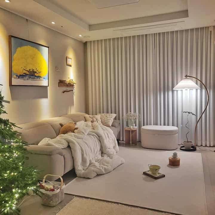 A white and natural tone living room featuring beige sofa, white curtains, cozy throw blanket, and round side table with a warm, inviting atmosphere
