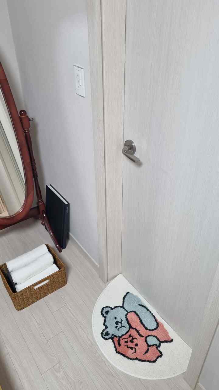 Natural-toned area in front of bathroom featuring a cute character bath mat, wooden full-length mirror, and rattan towel basket in a simple newlywed home interior