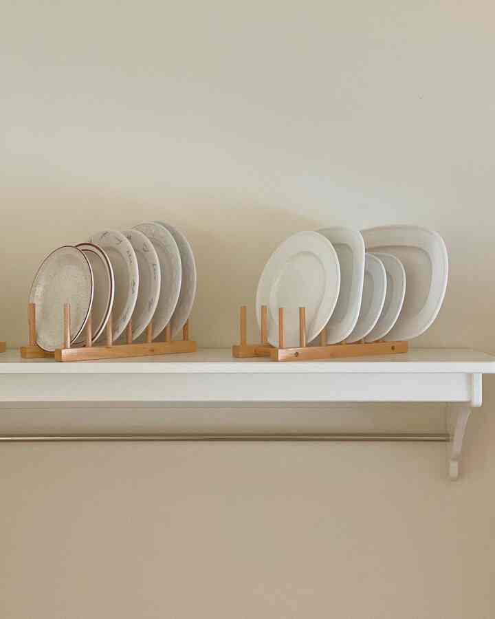 A kitchen plate storage space featuring wooden racks neatly holding various white plates on a white shelf against a light beige wall background