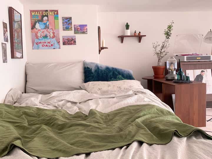 Bright beige-toned studio bedroom featuring wooden furniture, bed with plants and vintage posters creating a cozy atmosphere