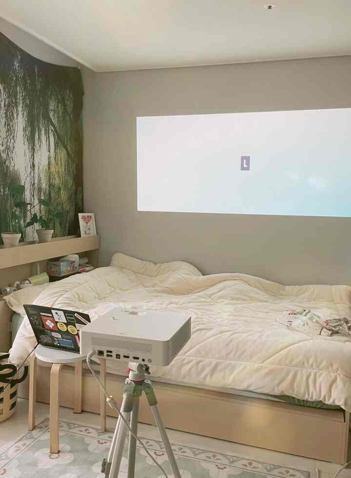 Soft beige and white toned bedroom featuring a bed, projector setup, and natural plants creating a cozy home cinema atmosphere