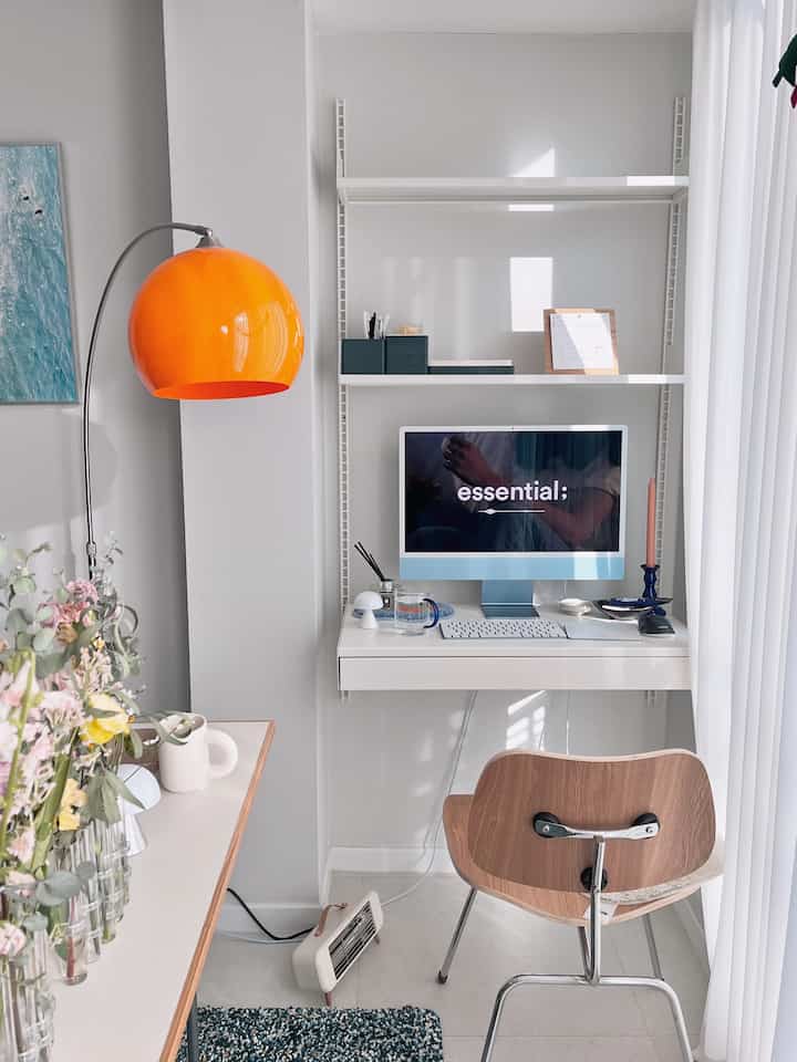 White-toned living room workspace featuring a compact desk and wooden chair with an orange floor lamp accent in a minimalist setting