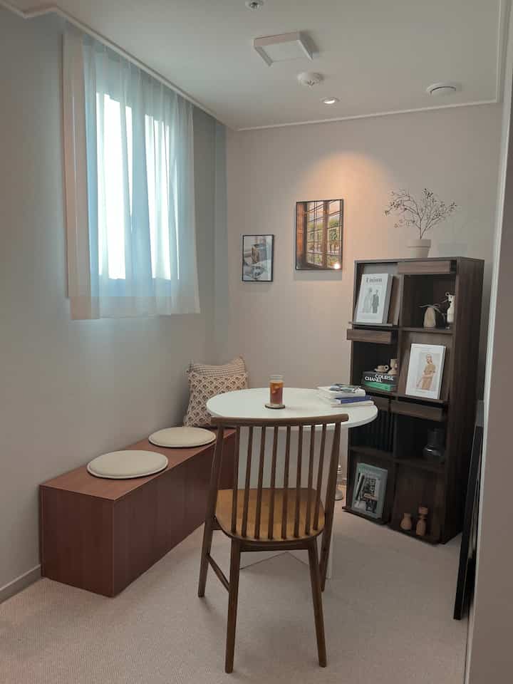 Natural-toned small home cafe featuring wooden chair and bench, white round table, and a bookshelf in a cozy interior