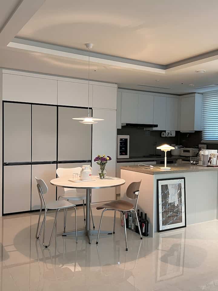 White and gray toned kitchen space featuring semi built-in refrigerator cabinet, round dining table with diverse chairs, and pendant lighting in a modern interior