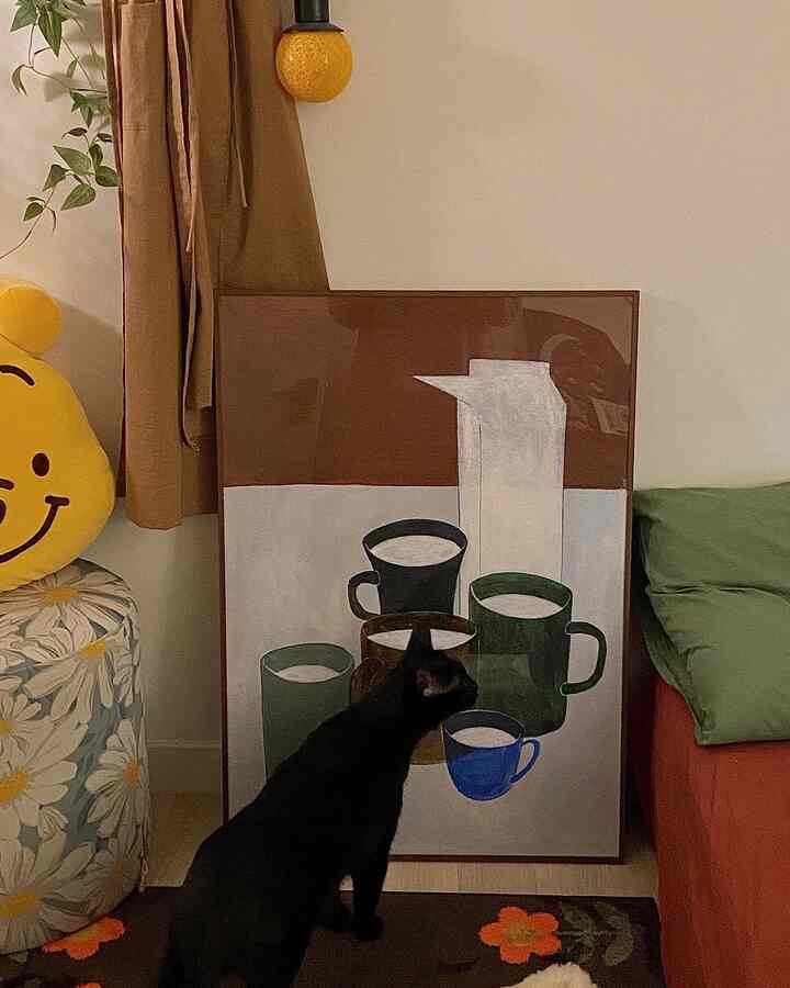 ブラウンとナチュラルグリーンの色調のリビング、黒猫と額装されたアート、花柄のカーペット、黄色いスマイリースツールが配された雰囲気のあるインテリア