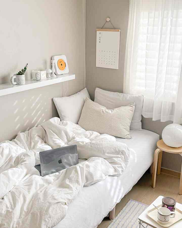 Beige-toned studio bedroom bathed in sunlight, featuring white bed and bedding with wooden stool creating a cozy atmosphere