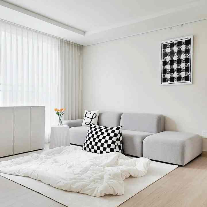 A white and gray toned living room featuring a modular sofa with checkered cushions, sheer curtains, and a rug with a white throw on the floor, presenting a clean space