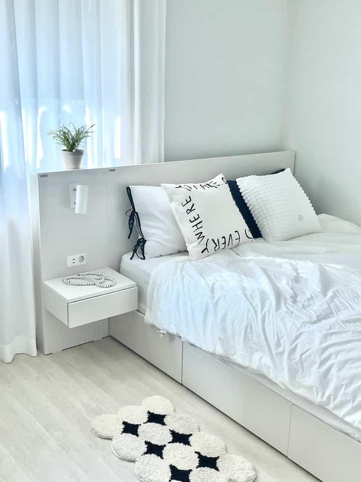Bright white-toned bedroom featuring simple bed with white bedding and a small plant on shelf, clean and minimalistic space
