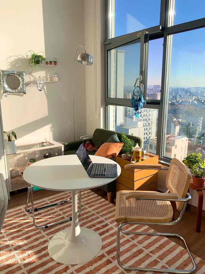 A white and brown toned loft apartment living room featuring a round table, cane chair, and green plants creating a cozy home office space