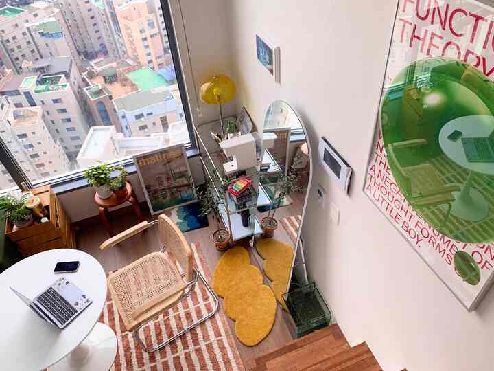 Bright natural-toned loft studio featuring a round table, rattan armchair, various plants, and rugs creating a cozy home office atmosphere