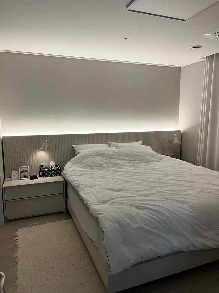 Soft white-toned bedroom featuring a large central bed, nightstand on the left, and curtains on the right creating a cozy atmosphere