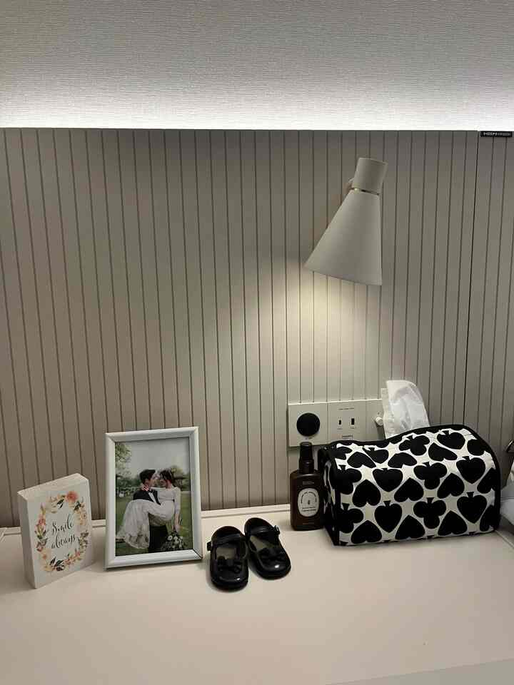 White and black toned bedroom with a wall-mounted lamp, black heart-patterned tissue box cover, and framed photo on a simple, elegant surface