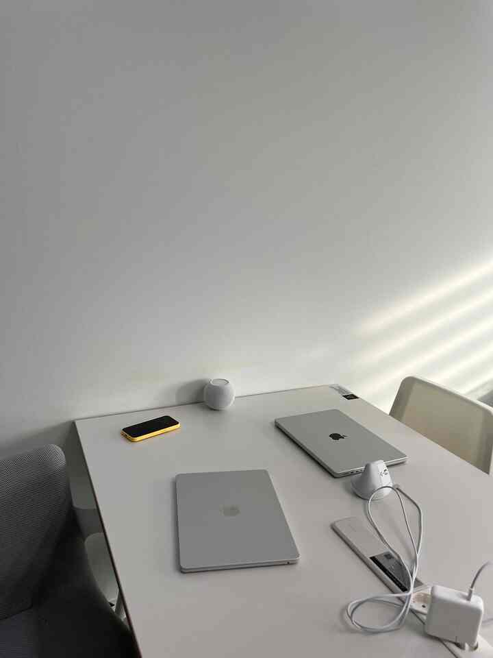 White-toned living room style home office featuring a large table with laptops and a Bluetooth speaker, a simple and clean workspace