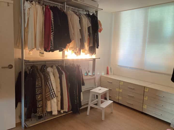 Beige and white toned walk-in closet featuring clothing racks on left, white stool in center, and long drawer cabinet on right for organized storage