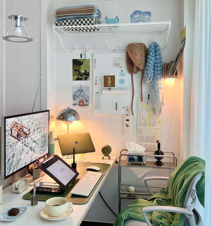 A white and green toned studio home office featuring a tidy desk, shelves, and postcards on the wall in a cozy workspace