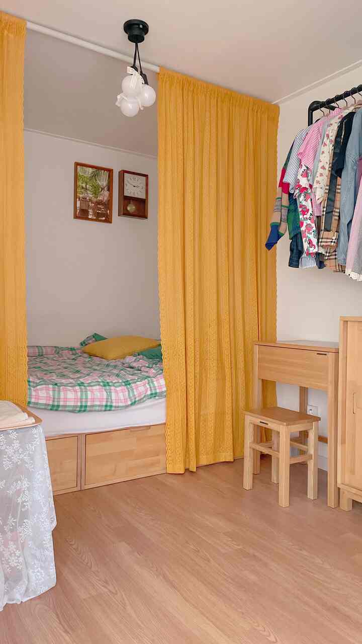 Cozy bedroom with warm wood tone flooring and yellow curtains, featuring a storage bed, clothes rack, and wooden desk