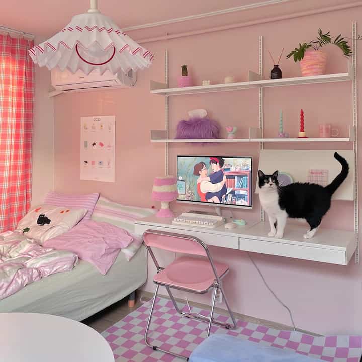 A compact bedroom with pink-toned walls and furnishings, featuring a desk with computer and a cat on top, creating a cozy atmosphere