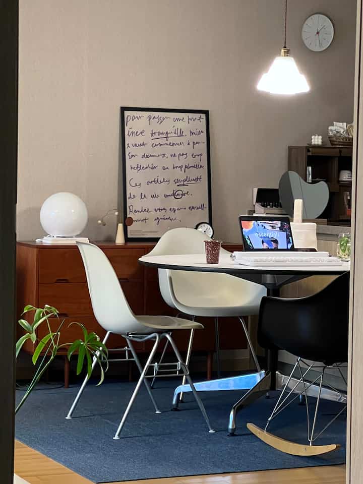 A modern and serene dining room with white and wood tones featuring a round dining table, Eames armchairs, and an Arne Jacobsen wall clock