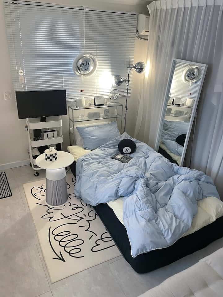 Sky blue toned bedroom with central bed, white blinds, side table, and full-length mirror in a cozy studio space